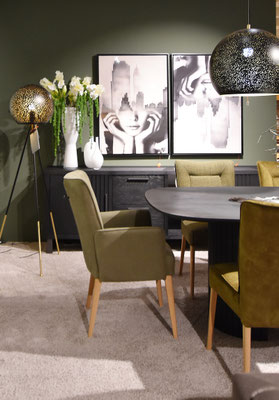 Ovaler, schwarzer Tisch mit olivgrünen Stühlen mit Holzbeinen, vor schwarzem Sideboard und Bildern.