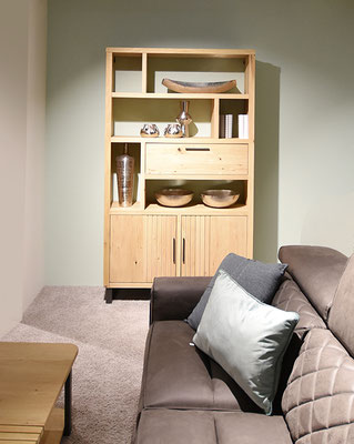 Braunes Ledersofa vor einem hohen, Holzregal mit Deko-Objekten in einem Raum mit hellem Teppich.
