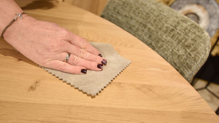 Hand wischt mit grünem Lappen über einen runden Holztisch; Stuhl im Hintergrund.