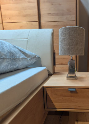 Detailansicht eines Holzbettes mit hellem Kopfteil, gemusterter Kissen und Nachttisch mit Lampe.