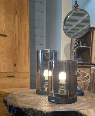 Auf einer Holzplatte stehen zwei Lampen aus dunklem Glas, dahinter steht eine Holzkommode.