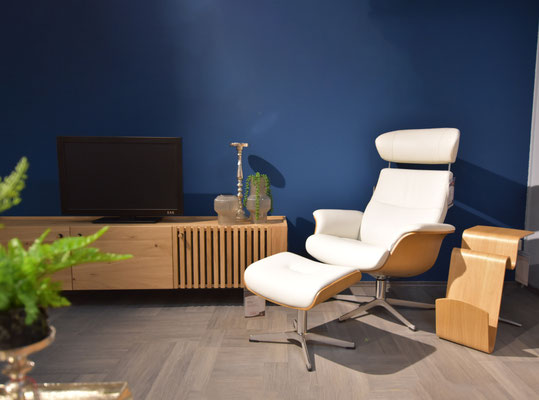  Weißer Ledersessel mit Ottomane vor einer dunkelblauen Wand, neben einem TV-Sideboard aus Holz.