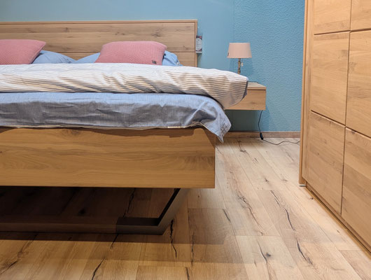 Holzbett und Kleiderschrank auf Holzboden vor einer blauen Wand mit Nachttischlampe.
