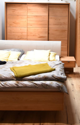 Holzbett mit weißem Kopfteil und grauer Bettwäsche, gelben Kissen und Decke vor einem Holzschrank.