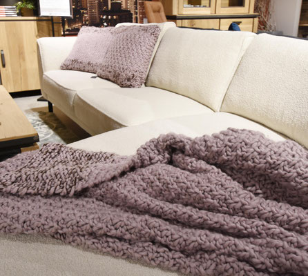 Weißes Sofa mit lila Strickkissen und einer dicken, grob gestrickten Decke in der gleichen Farbe.