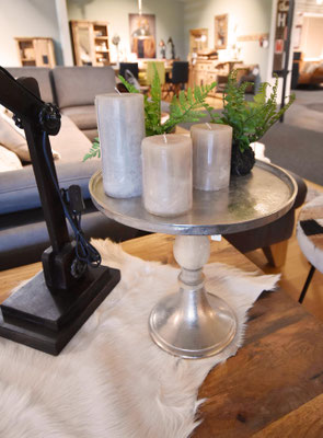 Runder Metalltisch mit drei Kerzen und Farn auf hellem Fell, davor eine schwarze Designerlampe.