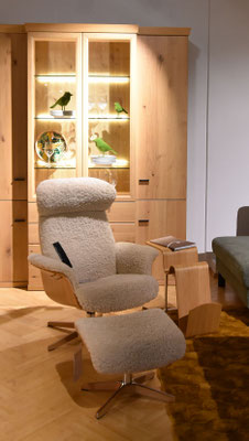 Ein cremefarbener Sessel mit Hocker vor einem beleuchteten Holzschrank mit Glasvitrine.