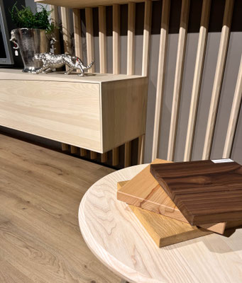 Holz- und Farbmuster auf einem hellen runden Beistelltisch vor einem Holzlamellen-Wandpaneel.