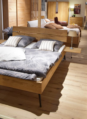 Eiche-Doppelbett mit geschwungenem Kopfteil, hellgrauer Bettwäsche und Nachttischlampe.