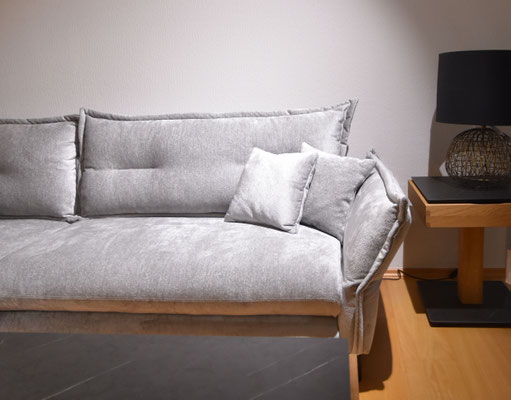 Helles graues Sofa mit weichen Kissen, daneben ein Beistelltisch mit schwarzer Tischlampe.