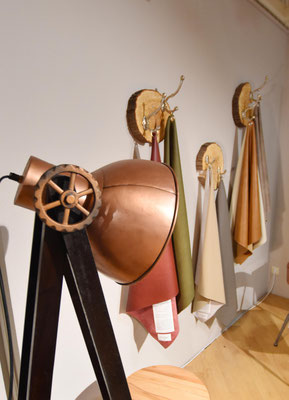 Kupferne Stehlampe im Industrial-Stil mit Holzzahnrad neben Wandhaken aus Holzscheiben mit Stoffen.