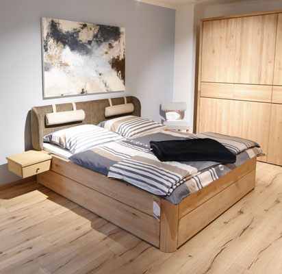 Holzbett mit Stoff bezogenem Kopfteil, gestreifter Bettwäsche und Decke mit Nachtisch und Schrank.