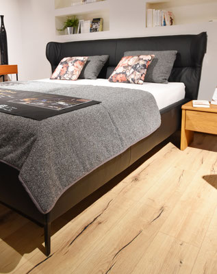 Schwarzes Doppelbett mit hohem Kopfteil, grauer Decke und Kissen sowie geblümten Kissen.