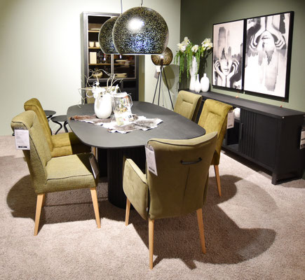 Ovaler, schwarzer Tisch mit olivgrünen Stühlen mit Holzbeinen, vor schwarzem Sideboard und Regal.
