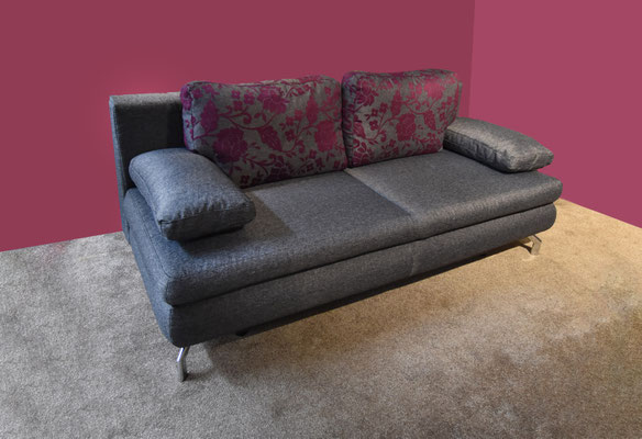 Ein dunkelgraues Zweisitzer-Sofa mit zwei violett-grau gemusterten Kissen vor einer pinken Wand.