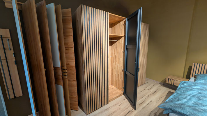 Offener heller Holzschrank mit Lamellenstruktur und schwarzer Tür in modernem Schlafzimmer.