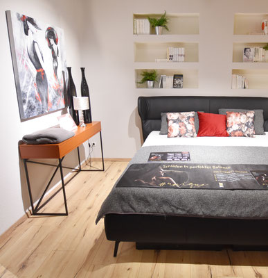 Schwarzes Doppelbett mit bunten Kissen neben einem Sideboard mit Lampe und anderen Gegenständen.
