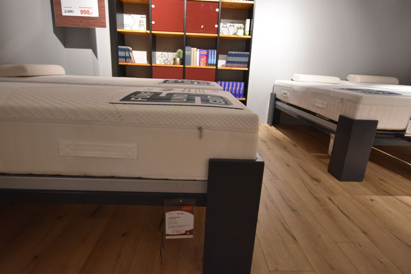 Weiße Matratze auf einem Bettrahmen mit Holzboden und Bücherregal im Hintergrund.