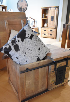 Rustikale Holztruhe im Wohnzimmer mit offenem Deckel und einem Fellkissen, dahinter ein Holzschrank.