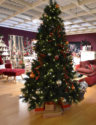 Großer Weihnachtsbaum, geschmückt mit Lichtern und goldenen Kugeln, rote Möbel im Hintergrund.