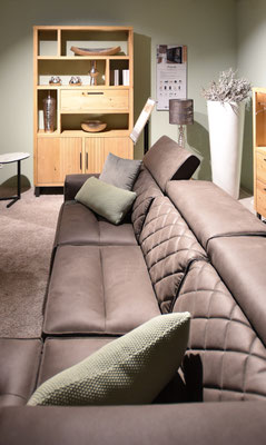 Braunes Sofa mit gestepptem Rücken und grünen Kissen dahinter ein Holzregal mit Fächern.