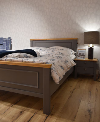 Dunkelgraues Holzbett mit hellbraunen Akzenten, gemusterter Bettwäsche, Nachttisch und Lampe.