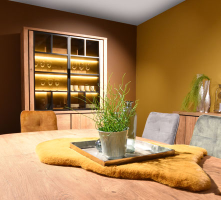 Esszimmer mit Holztisch, gelbem Fellläufer, Stühlen und beleuchteter Vitrine.