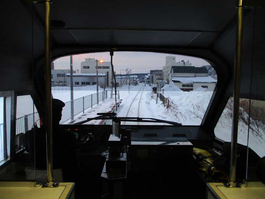 発車前、展望車最前にて運転士さんの後ろ姿と一緒に前方を見ます　ビルの影から朝日が昇ろうとしています