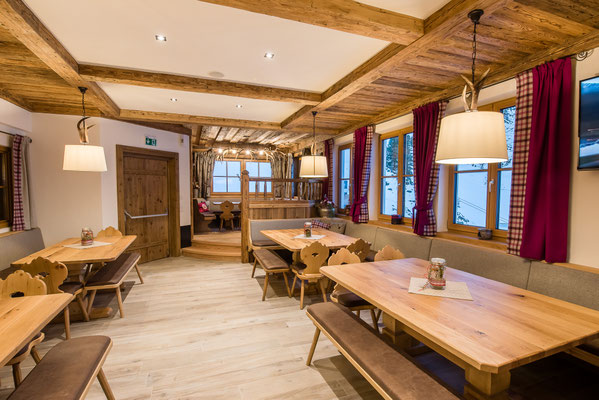 Frühstücksraum in der Skihütte Rettenstein mit Holztischen, Bänken, Stühlen, roten Vorhängen und sichtbaren Holzbalken an der Decke