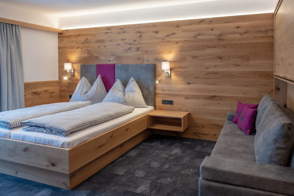 Schlafzimmer mit Holzwand, Doppelbett und grauem Sofa mit pinken Kissen
