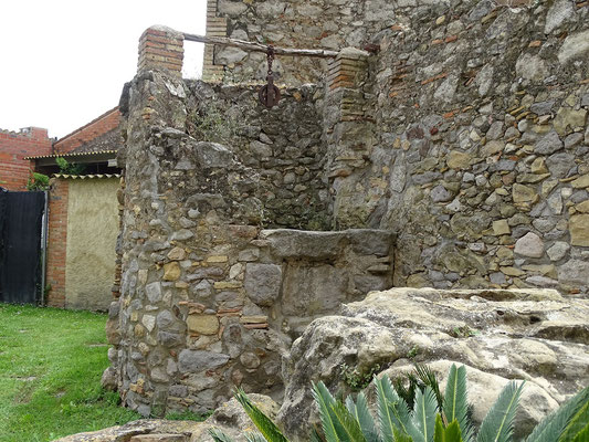 Bauerhof mit Ziehbrunnen vor der Kirche. Davor ein Felsen, der vom Wasser des früher hier vohandenen Sees ausgespült wurde