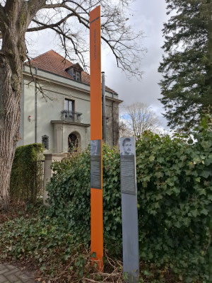 Eine orangefarbene und eine graue Gedenkstele vor einem Wohnhaus in Sacrow