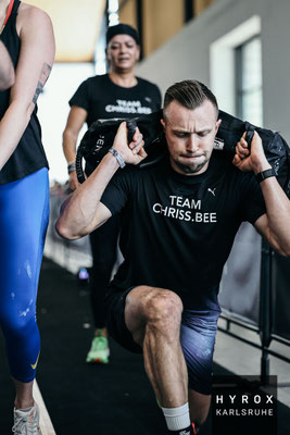 Christoph Wieloch macht Sandbag Lunges bei HYROX Karlsruhe 2025, Sabine Bell (Biene) ist im Hintergrund zu sehen.