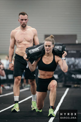Inga de Busman absolviert Sandbag Lunges beim HYROX Wettkampf in Barcelona, Christoph Wieloch folgt ihr im Hintergrund.
