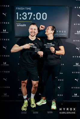 Sabine Bell (Bine) und Christoph Wieloch feiern das Finish bei HYROX Karlsruhe 2025 vor der Zielzeit 1:37:00 mit Finisher-Karten.