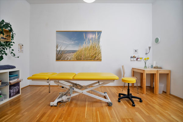 Ein ruhige Behandlungsraum mit einer Therapieliege und einem schönen Wandbild das einen Strand zeigt. Ein gelber Rollhocker steht vor dem Holztisch. Der Raum ist sauber und hell.