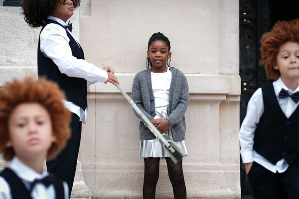 jeune file noire adossée à un mur pendant que des enfants jouent devant elle