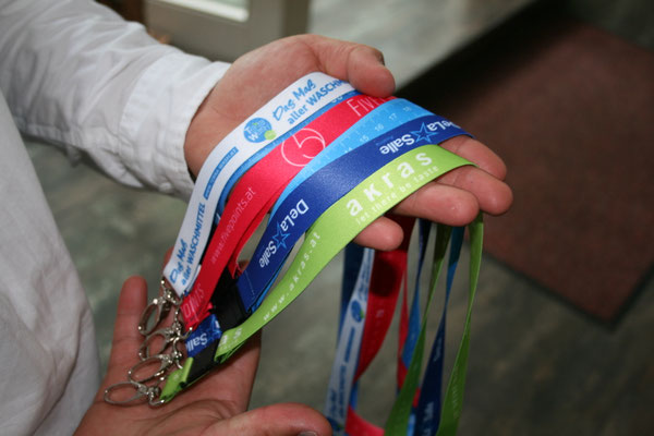 In zwei Händen werden verschiedene Lanyards gehalten. Darauf sind die Logos von Akras, Fivepoints und der Schule de la Salle erkennbar.
