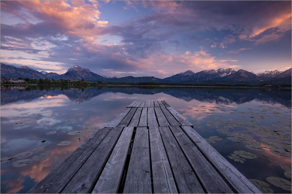 Der Hopfensee, gelegen in der malerischen Region Bayerisch Schwaben, ist ein idyllisches Naturjuwel. Umgeben von sanften Hügeln und der imposanten Kulisse der Bayerischen Alpen, bietet der See ein perfektes Panorama für Fotografie-Enthusiasten