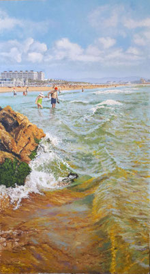 Playa de la Patacona con bañistas en el agua, rocas en primer plano y edificios residenciales al fondo en un día soleado.
