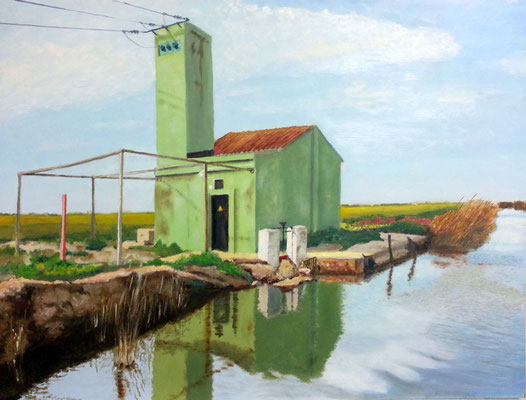 Edificio verde junto a un canal, con torre estrecha, reflejo en el agua y paisaje agrícola alrededor.