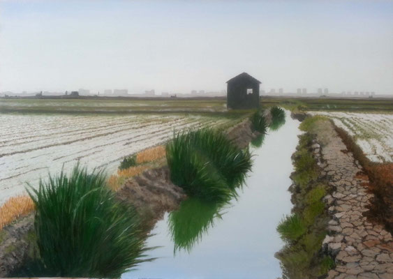 Acequia con orillas de barro seco cuarteado, vegetación alta, caseta oscura al fondo y edificios de El Perellonet en el horizonte.