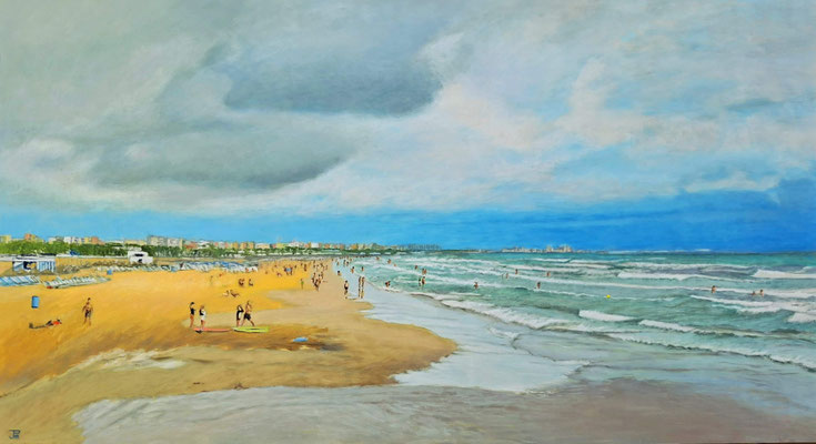 Playa amplia con arena dorada, gente paseando junto a la orilla, mar con oleaje y cielo con nubes densas que se abren hacia zonas de azul luminoso.