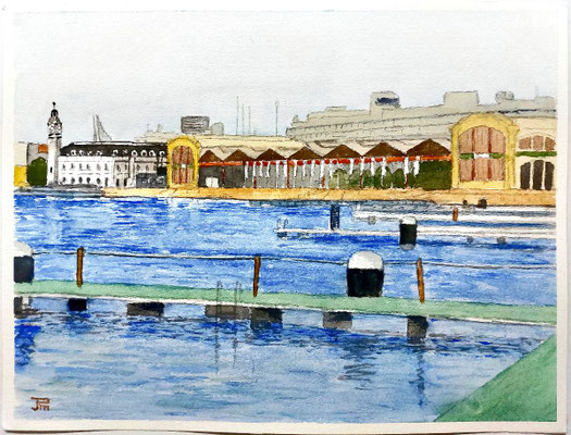 Vista del puerto con agua en primer plano, postes y amarres, edificios portuarios con tejados triangulares y una torre al fondo bajo cielo gris.