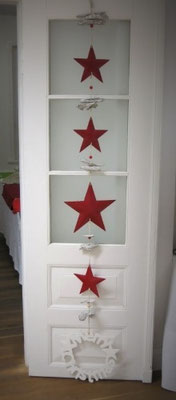 Lange, rote Girlande mit roten Metallsternen und weißem "Merry Christmas" Schild an eine weiße Tür gehängt.