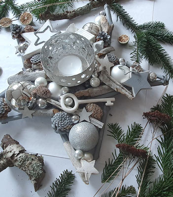 Adventsgesteck in sternform mit einem weiß-silbernen Metallwindlicht, grauen Holzsternen und Schlüssel.