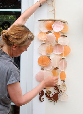Ina hält ein Capiz Windspiel aus apricot- und orangefarbenen Muschelscheiben mit Edelrost Seepferdchen. vor eine weiße Hauswand