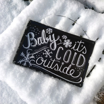 Metallschild auf Schnee: "Baby it`s cold outside" heißt der weiße Schriftzug auf schwarzem Untergrund.