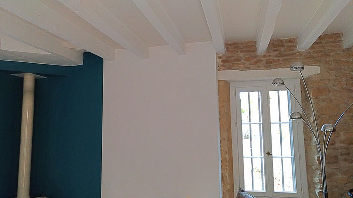 Rénovation d'une maison en pierre. Grand salon bleu et blanc avec mur en pierre apparente. Plafond et poutres peints en blanc et beige.