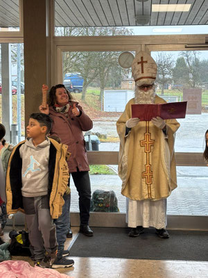 Der Nikolaus liest etwas aus seinem Roten Buch vor. Im Hintergrund sieht man die Schulseretärin mit einem Geschenk in der Hand. Im Vordergrund ist ein Schüler der ersten Klasse zu sehen.